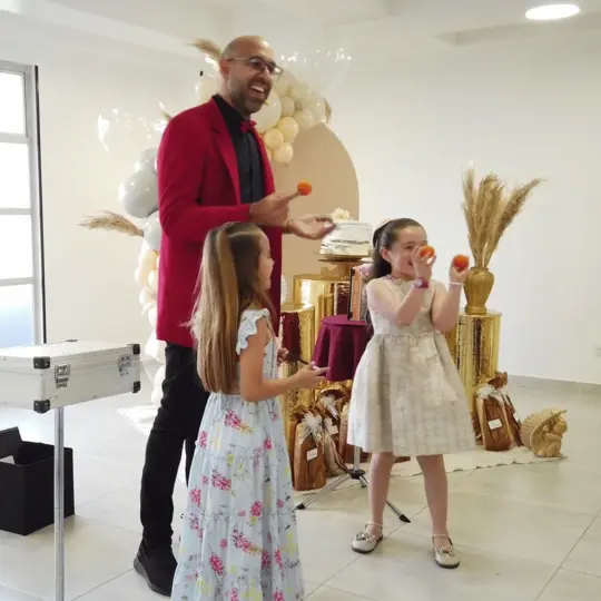 Mago profesional en chaqueta roja riendo mientras realiza un truco de magia interactivo con dos niñas asistentes que sostienen pelotas naranjas en una fiesta infantil decorada en Bogotá.