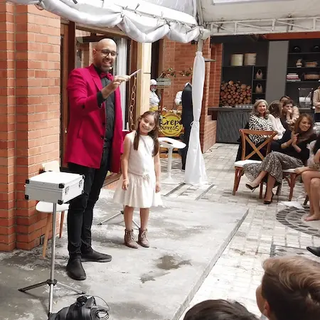 Mago profesional realizando un truco de magia con una niña asistente en un show de magia familiar al aire libre en Bogotá. Espectáculo de entretenimiento para eventos infantiles y reuniones familiares.